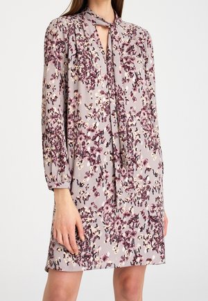 Frau trägt ein helllila Blumenkleid mit langen Ärmeln und einem gebundenen Ausschnitt, steht vor einem einfarbigen weißen Hintergrund.