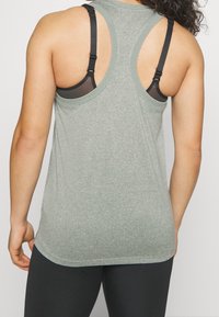 Top atletico grigio chiaro con design a schiena scoperta. Presenta spalline nere e un tessuto morbido e texture. L'orlo curva leggermente nella parte inferiore.