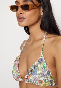 Top de bikini com padrão floral e bordas com babados; as cores incluem rosa, laranja, amarelo e verde. Acompanhado de óculos de sol retangulares marrons.