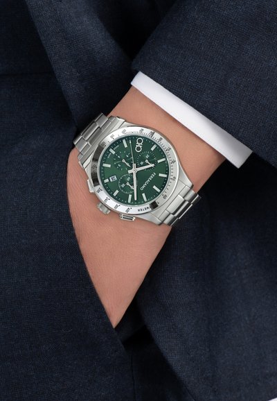 Încheietură cu ceas cronograf Ferragamo din metal argintiu, cu cadran verde, marcaje albe și fereastră pentru dată, pe un fundal de material pentru costum de culoare albastru închis.