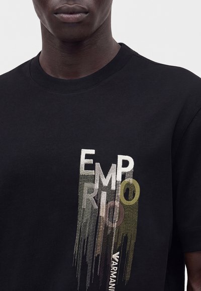 Άνδρας φοράει μαύρο T-shirt με πολύχρωμα κεντημένα γράμματα που σχηματίζουν "EMPORIO" και "ARMANI" στο στήθος.