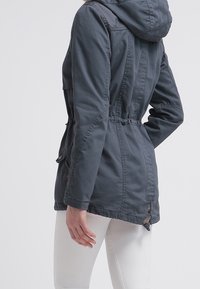 Chaqueta gris con capucha y cintura fruncida, con cierre de botones y bolsillos, hecha de un tejido texturizado. Pantalones blancos visibles debajo.
