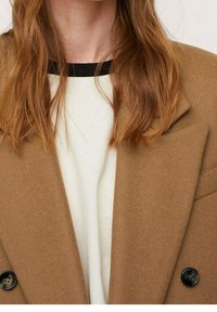 Brun uld blazer med spidse lapeller, dobbeltradet design og sorte knapper, parret med en hvid top med en sort udskæring.