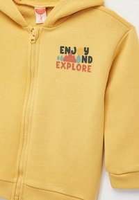 Gelbes Zip-Up-Hoodie mit Kängurutasche und Kapuze. Siebdrucktext "GENIEßEN UND ENTDECKEN" mit Grafiken von Bergen und Bäumen.