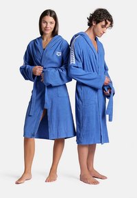 Blauwe katoenen badjassen met capuchon, voorzien van opgestikte zakken en een bindceintuur. Eén heeft een logo, de ander een contrasterende zijstrook.