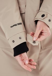 Vedenpitävä beige takki, jossa on W-PRO 10000 -branding, jossa on nappikiinnitys hihassa ja musta ribbivuori. Kädentiet säädetään.