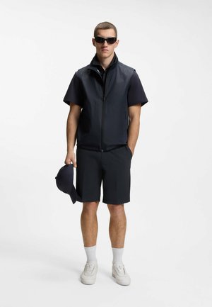 Mężczyzna noszący czarne okulary przeciwsłoneczne, czarną koszulkę z krótkim rękawem, czarną kamizelkę na zamek, czarne spodenki, białe skarpetki, białe trampki, trzymający czarną czapkę.