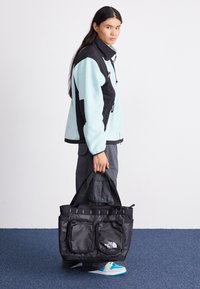 Saco tote preto com dois bolsos exteriores e logo, feito de tecido durável. Usado por um indivíduo com uma jaqueta de fleece azul claro e calças cinzas.
