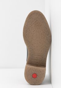 Semelle d'une chaussure Kickers marron avec une semelle texturée antidérapante et un détail circulaire rouge près du talon, posée sur un fond blanc.