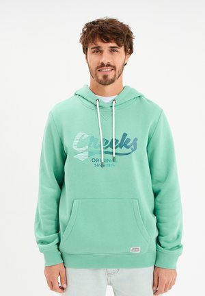 Mann mit kurzen lockigen Haaren und Bart, der einen mintgrünen Hoodie mit dem Schriftzug „Creeks Original Since 1974“ und hellblaue Jeans trägt, steht vor weißem Hintergrund.