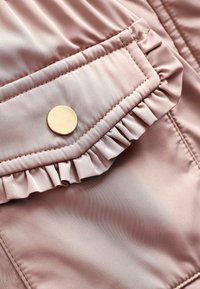 Tissu satin rose avec une poche à rabat froncée maintenue par un bouton rond en métal doré sur un vêtement.