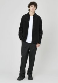 Chemise boutonnée en velours côtelé noir sur un t-shirt blanc, associée à un pantalon noir et des chaussures noires. Le modèle se tient contre un fond neutre.
