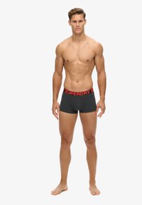 Boxer shorts negros con un logo rojo de "SUPERDRY" en la cinturilla. La tela es suave, ajustada y sin costuras, diseñada para la comodidad.
