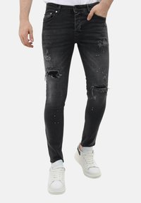 Zwarte distressed skinny jeans met rafelige scheuren en witte spikkels. De stof heeft een soepele textuur; gecombineerd met witte sneakers.
