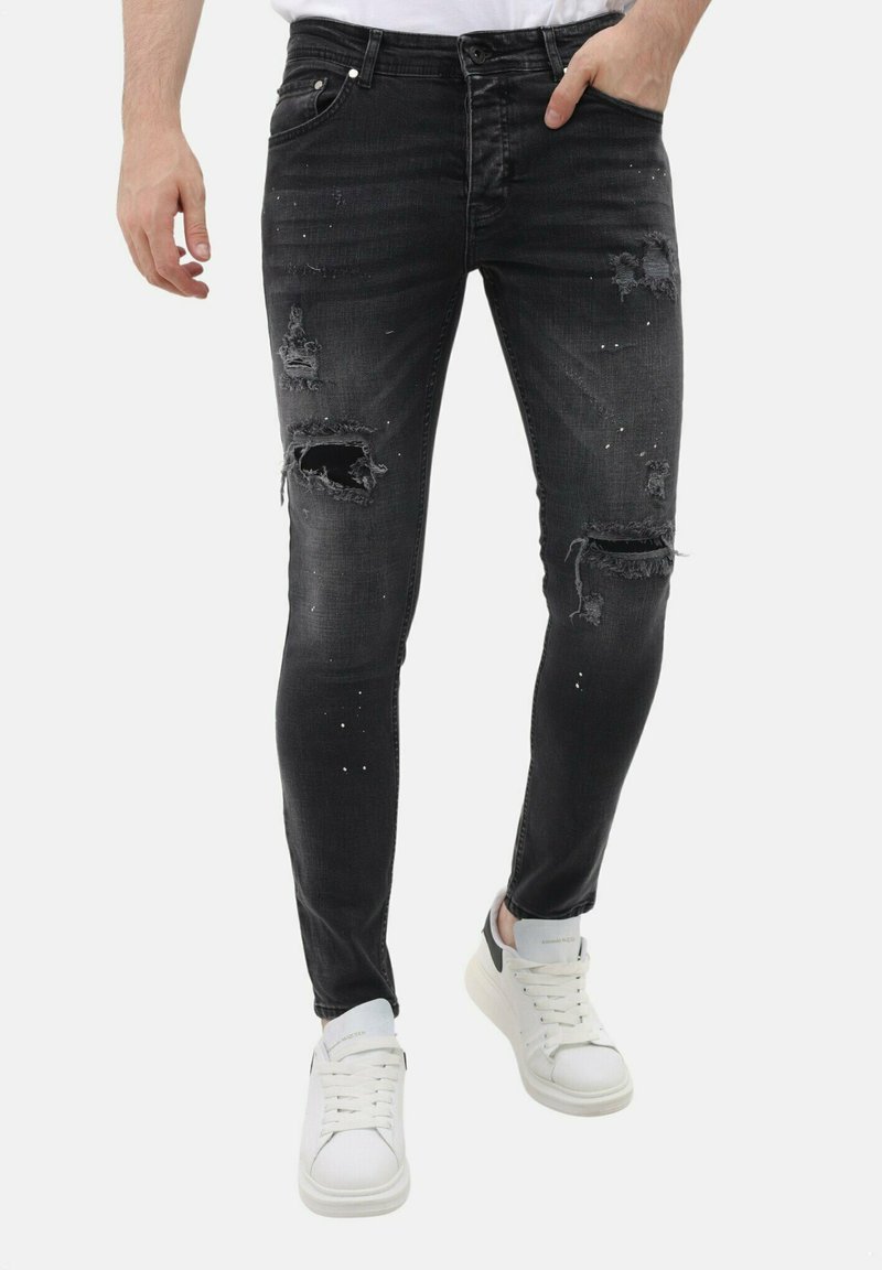 Zwarte distressed skinny jeans met rafelige scheuren en witte spikkels. De stof heeft een soepele textuur; gecombineerd met witte sneakers.