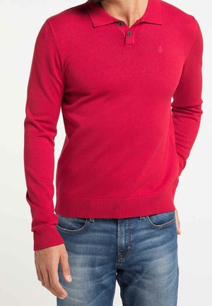 Man wearing bright red long-sleeve polo shirt and blue jeans, standing with one hand in pocket and one hand relaxed by side.