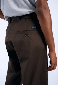Braune Baumwollhose mit einer Gesäßtasche und einem schwarzen Etikett. Die Hose hat eine gerade Passform und sichtbare Nahtdetails.