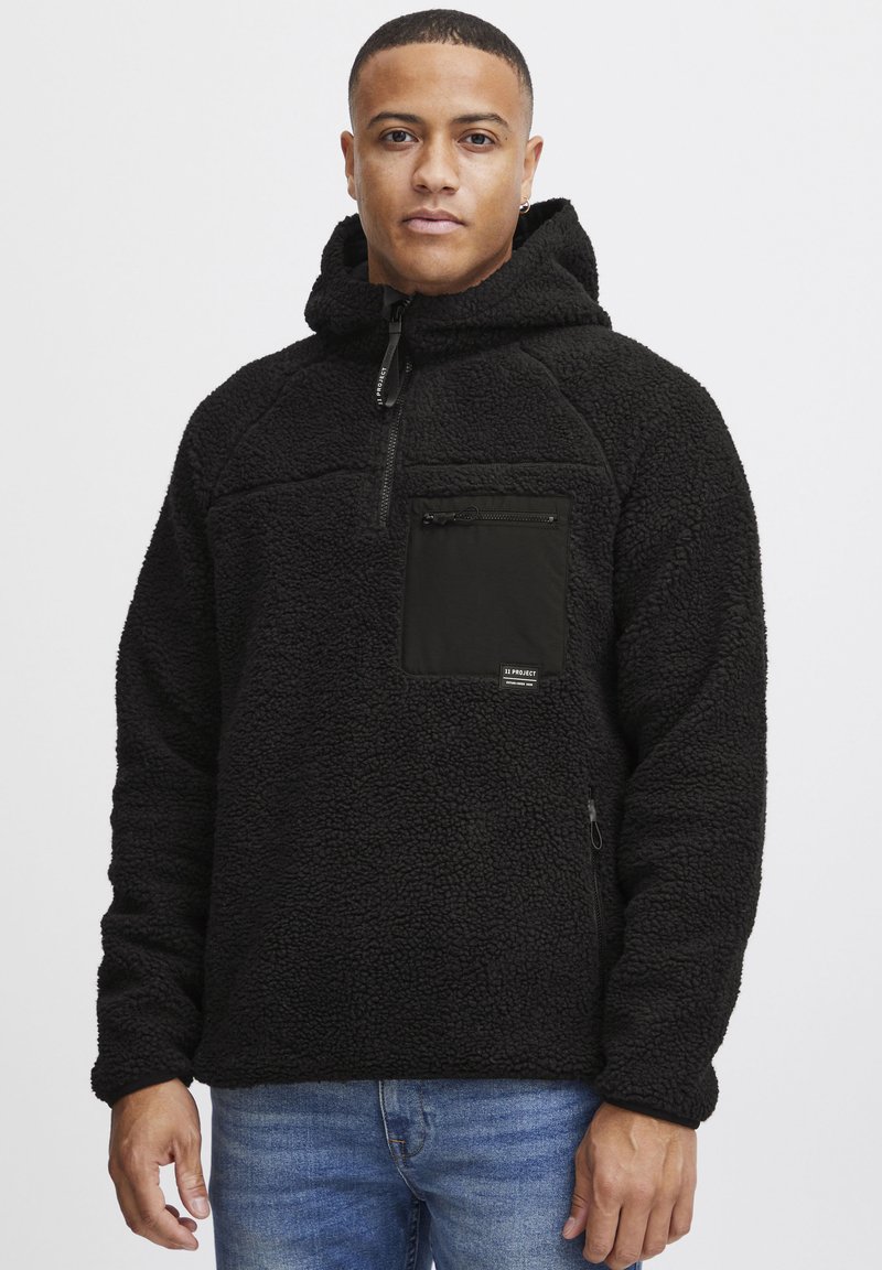 Svart fleece genser med glidelås i brystlommen, teksturert overflate og justerbar hette. Har sidelommer med glidelås og kontrastfargede detaljer.