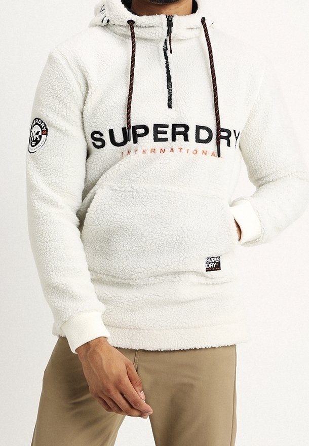 Homme portant un sweat à capuche en polaire blanche Superdry avec poche avant et pantalon beige, main partiellement dans la poche et visible sur fond blanc.