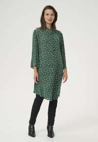 Robe chemise verte à pois, avec un devant à boutons, des manches 3/4 et un ourlet arrondi. Portée sur un pantalon noir et des bottines noires.
