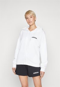 Fehér cipzáras kapucnis pulóver, állítható kapucnival, fekete Adidas logóval a mellkason. Fekete sportos rövidnadrággal párosítva, minimalista dizájnnal.