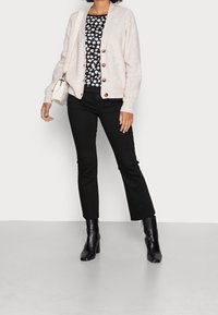 Cardigan beige in maglia con bottoni in legno, indossato sopra un top floreale nero, abbinato a jeans neri a zampa d'elefante e stivaletti neri con tacco.