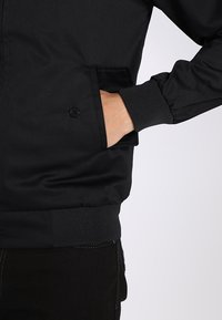 Giacca bomber nera, tessuto testurizzato, polsini e vita a coste, con una tasca laterale e dettaglio a bottone, esposta su uno sfondo neutro.