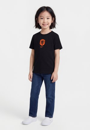 Lachend kind staand, gekleed in een zwart T-shirt met een oranje leeuw-embleem, blauwe jeans en witte instapschoenen tegen een effen witte achtergrond.