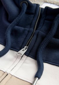 Námornícka a béžová mikina s kontrastným zipsom, textúrovanými šnúrkami na stiahnutie a dizajnom s panely, vyrobená z mäkkej látky s viditeľnými švami.