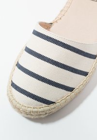 Sandały espadrille z beżową cholewką z kanwy, z niebieskimi paskami. Wykonana z plecionej juty podeszwa z haftowanymi brzegami dla dodatkowej tekstury.