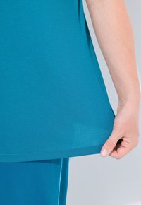 Tyrkysový top s krátkým rukávem vyrobený z měkkého, texturovaného materiálu. Má zakřivený lem a přiléhavý střih, na fotografii mírně tažený dole.