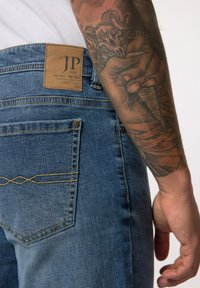 Los jeans de mezclilla azul cuentan con un parche de cuero con la etiqueta "JP 1880" en la cintura, costuras amarillas y bolsillos traseros con costuras decorativas.