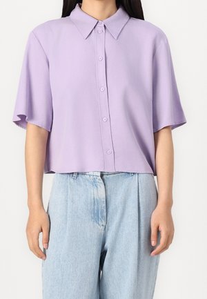 Femme portant une chemise courte lavande à boutons avec manches courtes et un jean ample bleu clair taille haute sur fond blanc.
