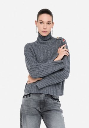 Maglione grigio a coste con collo alto e motivo a rombi, con un accento sulla spalla decorato da un bottone; indossato con jeans grigi.