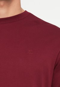 Burgundia värvi ümar kaelusega T-särk, mis on valmistatud pehmest puuvillasegust, ning millel on rinnal toon-toonis brodeeritud logo ja lühikesed varrukad.