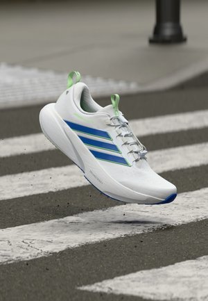 Witte sportschoen met groene accenten en blauwe strepen, bovenwerk van mesh, vetersluiting en gevoerde zool, afgebeeld in de lucht boven een zebrapad.