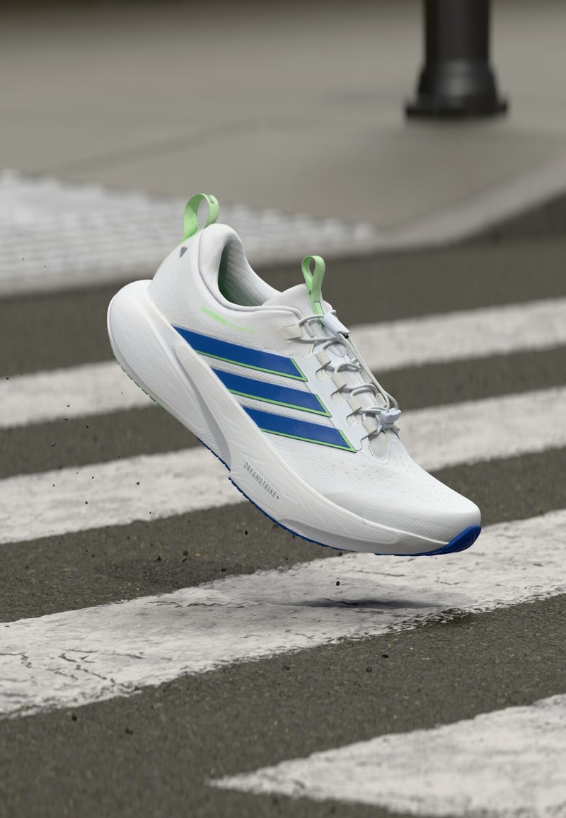 Chaussure de sport blanche avec accents verts et rayures bleues, empeigne en mesh, design à lacets et semelle rembourrée, représentée en plein saut au-dessus d'un passage piéton.