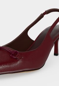 Chaussure à talon haut slingback en cuir rouge avec un petit détail décoratif en boucle à l’avant et un talon fin de hauteur moyenne.
