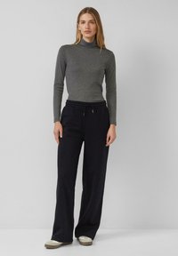 Kobieta w szarym golfie, czarnych szerokich spodniach z sznurkiem i beżowych sneakersach stoi na tle jasnoszarego, gładkiego tła.