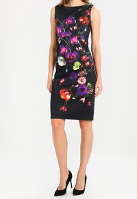 Černé bezramínkové šaty ke kolenům s multicolor květinovým potiskem, které modelka nosí s černými vysokými podpatky, stojí na bílém pozadí.