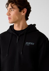 Sweat à capuche zippé noir en tissu doux, avec un logo avant en bleu sarcelle, des cordons réglables et une coupe décontractée.