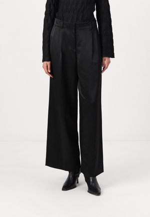 Femme portant un pantalon large en satin noir, un haut noir texturé à manches longues et des bottines noires à bout pointu, debout sur un sol blanc uni.