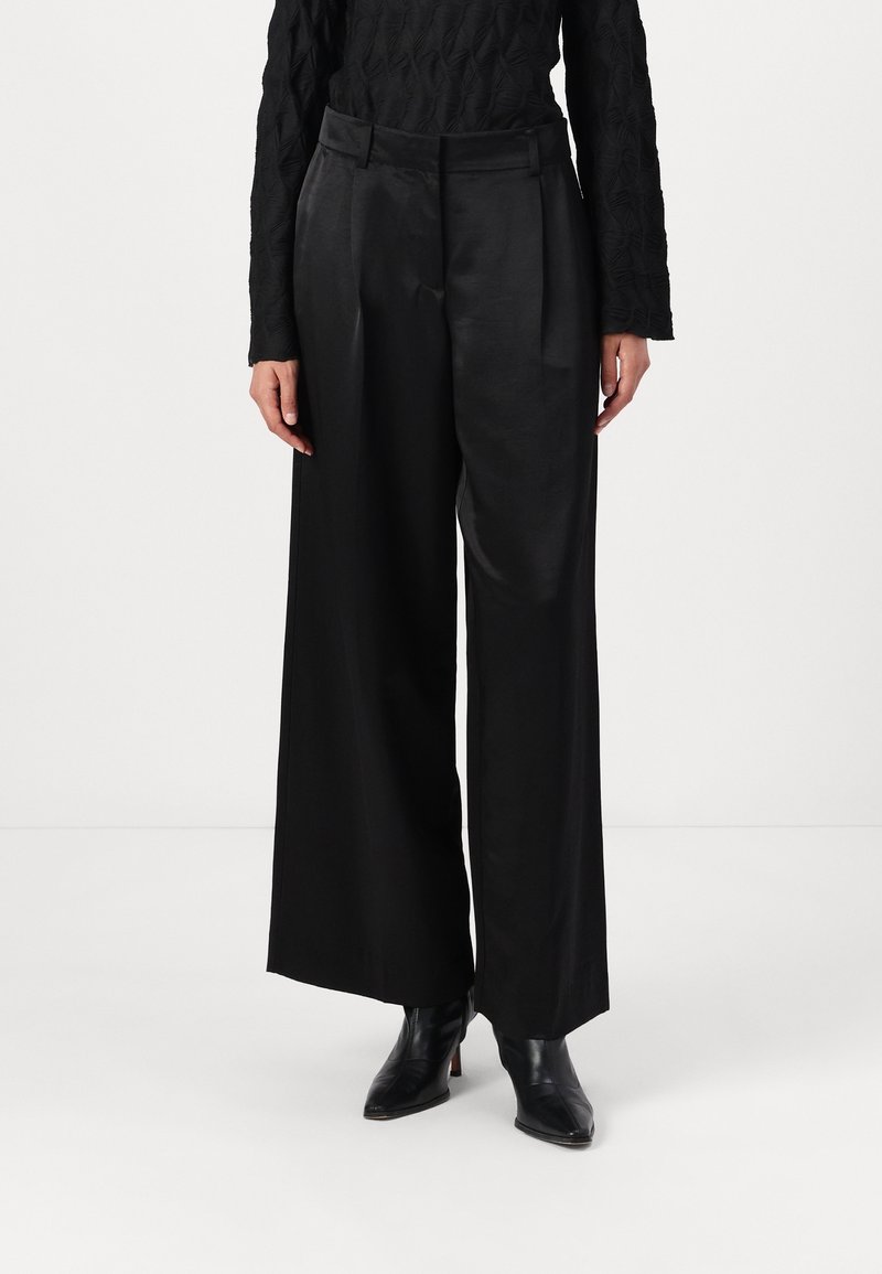 Femme portant un pantalon large en satin noir, un haut noir texturé à manches longues et des bottines noires à bout pointu, debout sur un sol blanc uni.