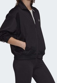 Woman wearing a black zip-up jacket with elastic cuffs and hem, hands in pockets, paired with black pants and a light gray shirt underneath.