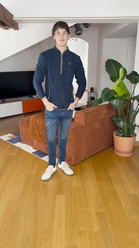 Veste zippée marine, jeans bleu foncé et baskets blanches. L'arrière-plan comporte un canapé couleur terre cuite, une plante verte et un parquet en bois.