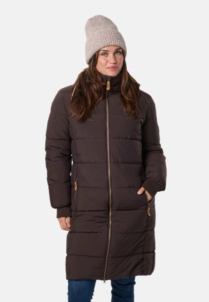 Une femme présente un long manteau matelassé marron foncé à fermeture éclair sur le devant. Elle porte un bonnet en maille beige et un jean bleu sur un fond uni.