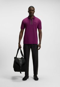 Herrkortärmad polo i livlig lila, med en dragkedjekrage och svart logotyp. Kombinerad med svarta byxor och en texturerad svart duffelväska.