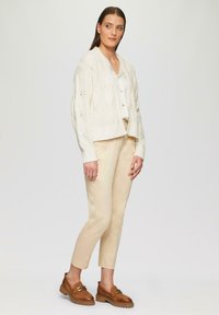 Cremefarbener, gestrickter Cardigan mit strukturierten Mustern, weiße Bluse, beige Hose und braune Loafers mit goldenen Hardware-Akzenten.