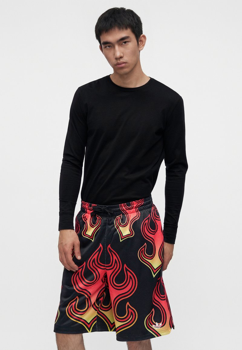 Giovane uomo con maglietta nera a maniche lunghe e pantaloncini neri con motivi di fiamme rosse e gialle, in piedi davanti a uno sfondo semplice.