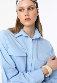 Jasnoniebieska koszula zapinana na guziki z dwiema kieszeniami na piersi, długimi rękawami i gładką teksturą. Modelka nosi na nadgarstku dużą kremową bransoletkę.